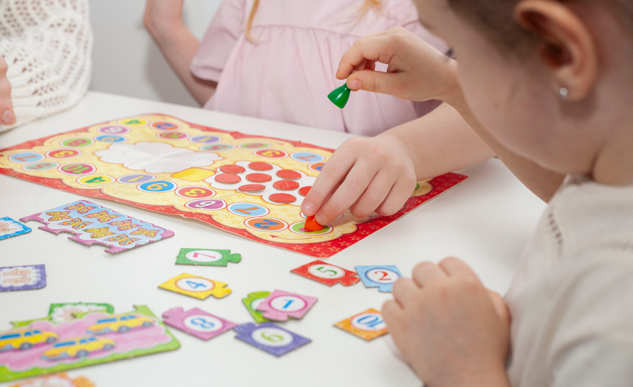 A child's hands placing a colored counter onto a numbered board game at a white table.